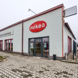 Fasada budynku z dużym czerwonym logo 'mikea' nad szklanymi drzwiami wejściowymi; obok widoczny szyld 'salon sprzedaży stolarki budowlanej' i parking z kostki brukowej.
