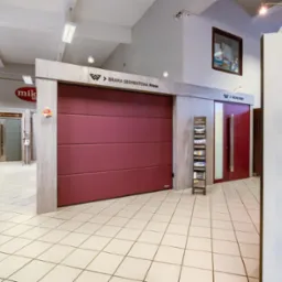 Ekspozycja w salonie: bordowa brama segmentowa Prime marki Wiśniowski, obok metalowy stojak z katalogami, w tle drzwi CREO oraz ekspozytor z klamkami Italian Design infinity line.