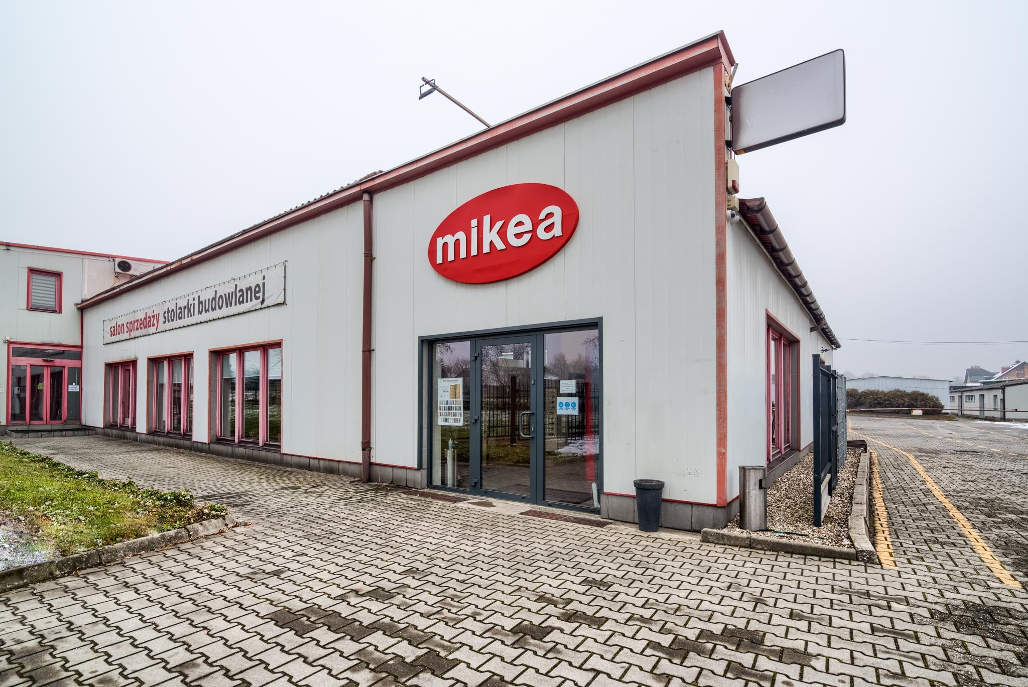 Fasada budynku z dużym czerwonym logo 'mikea' nad szklanymi drzwiami wejściowymi; obok widoczny szyld 'salon sprzedaży stolarki budowlanej' i parking z kostki brukowej.
