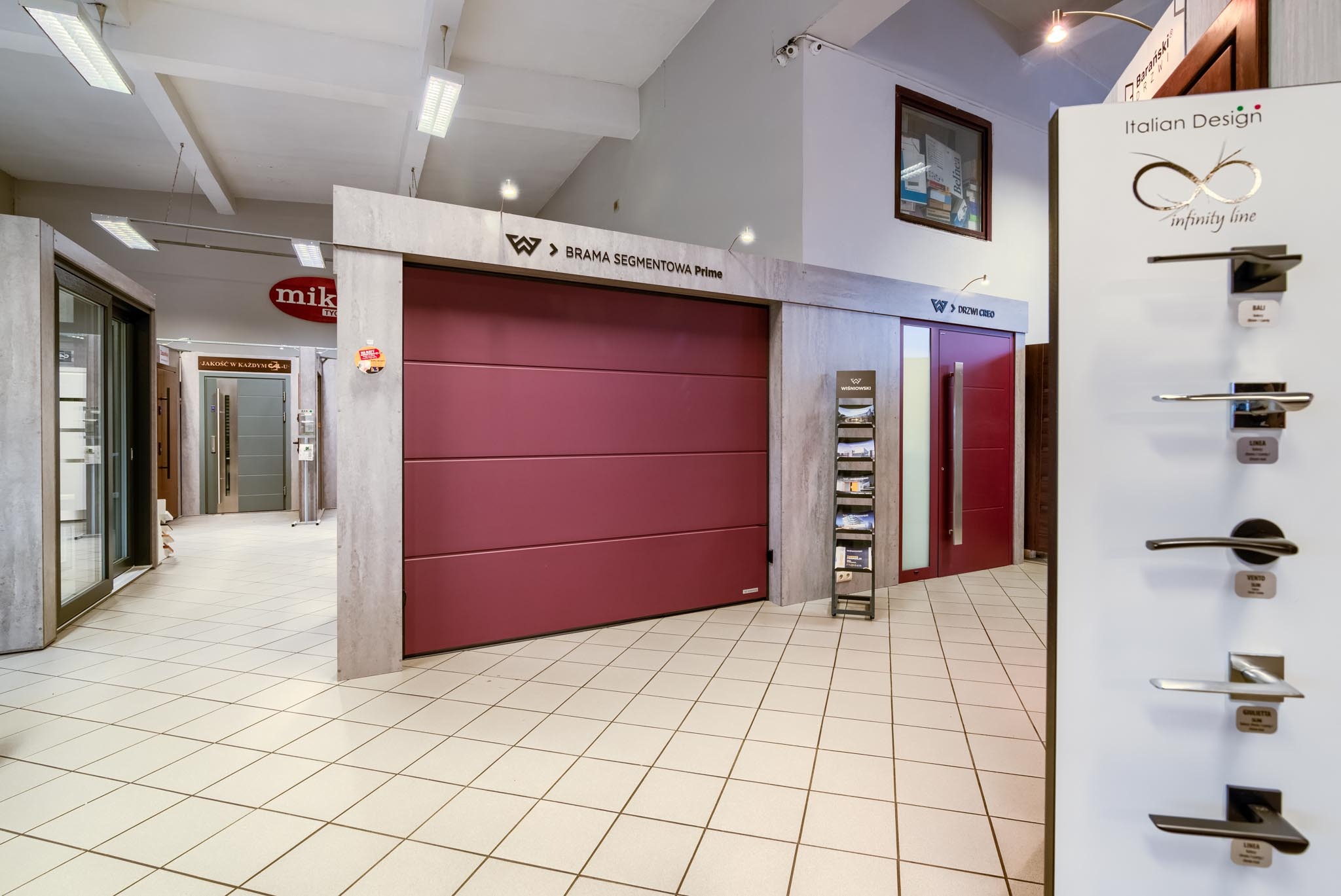 Ekspozycja w salonie: bordowa brama segmentowa Prime marki Wiśniowski, obok metalowy stojak z katalogami, w tle drzwi CREO oraz ekspozytor z klamkami Italian Design infinity line.