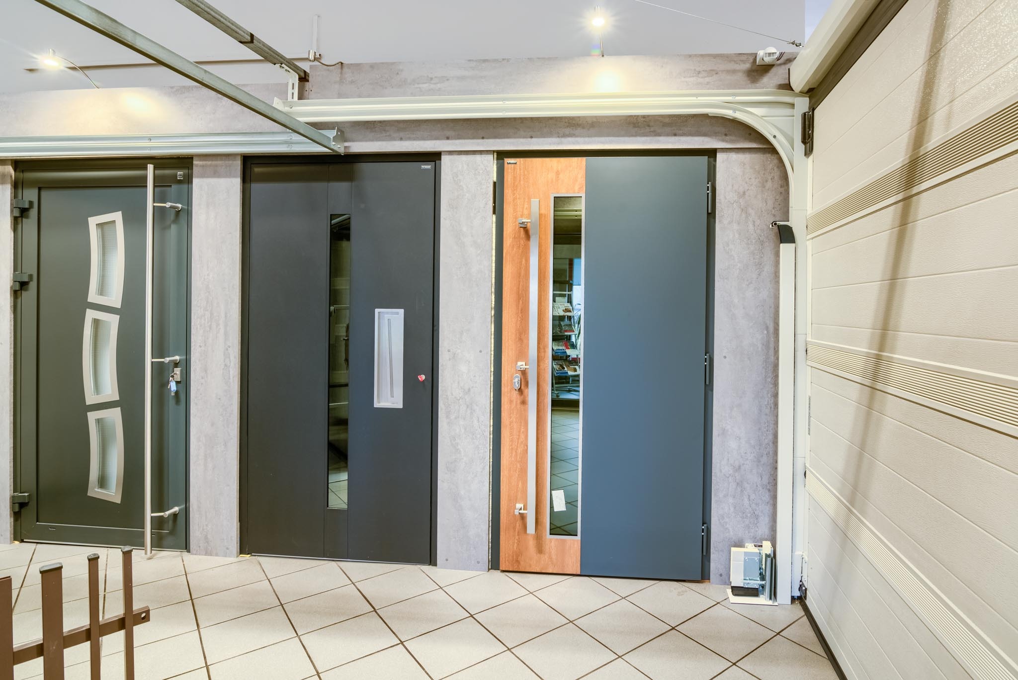 Ekspozycja w salonie: trzy różne modele drzwi wejściowych w odcieniach szarości i brązu, obok fragmentu białej bramy garażowej panelowej z poziomymi przetłoczeniami, na podłodze jasne płytki w kratkę.