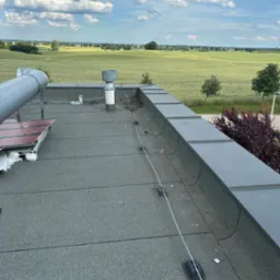 Widok z góry na płaski dach pokryty papą z widocznym kominem wentylacyjnym, rurą wentylacyjną oraz elementami instalacji odgromowej, w tle rozległe pole i pochmurne niebo.