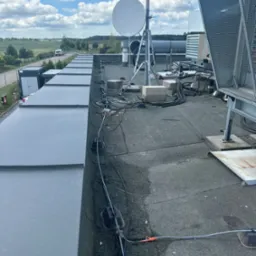 Płaski dach pokryty papą z widocznymi instalacjami telekomunikacyjnymi: anteny satelitarne, kable, oraz elementy mocujące. W tle widoczne budynki kontenerowe i krajobraz z polami i lasami...