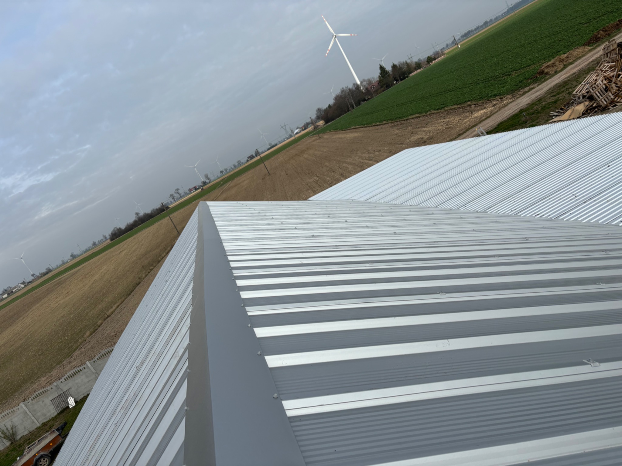 Nowy, blaszany dach trapezowy w kolorze srebrnym, widok z góry na tle pól uprawnych i turbin wiatrowych w oddali.