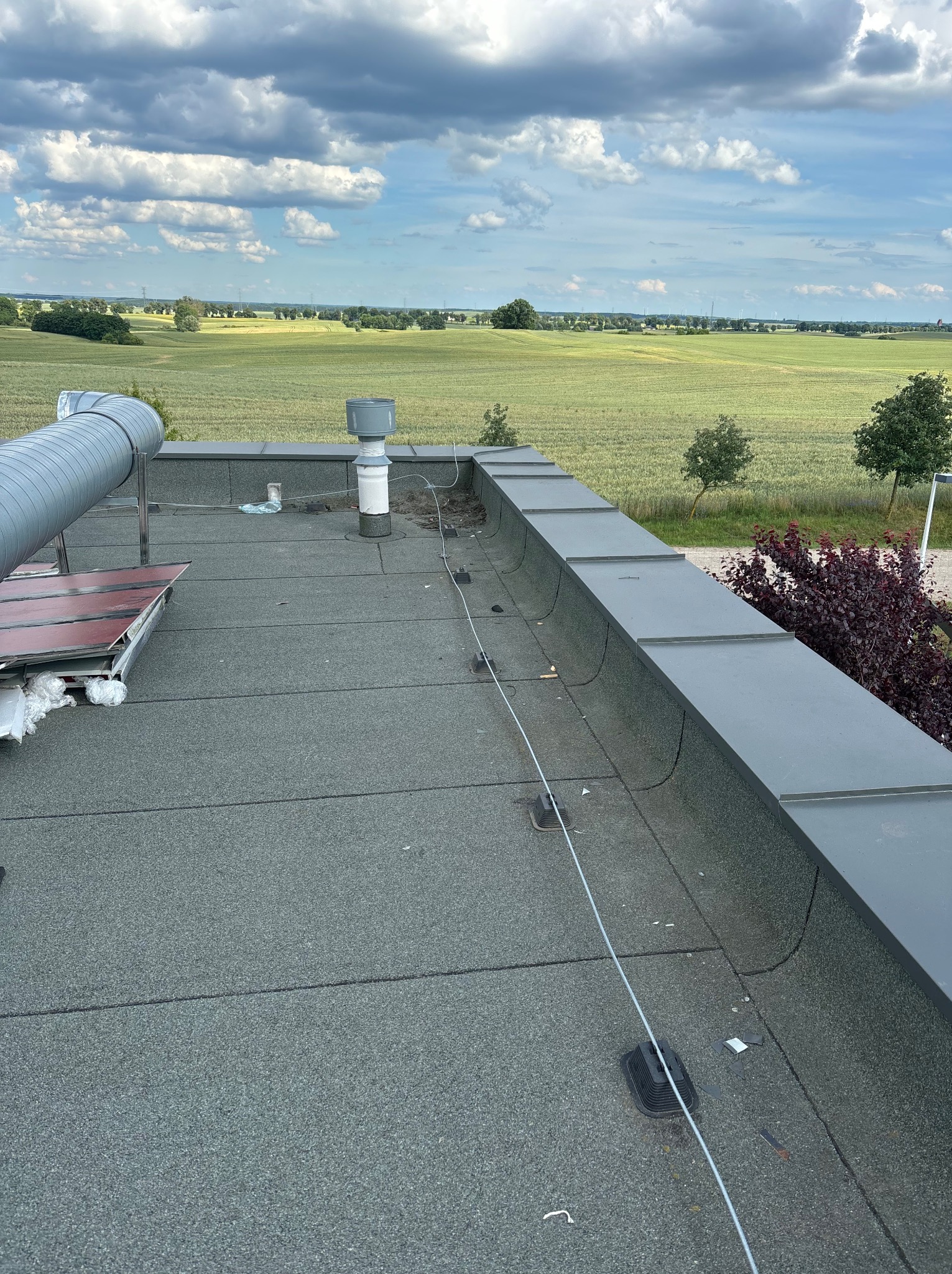 Widok z góry na płaski dach pokryty papą z widocznym kominem wentylacyjnym, rurą wentylacyjną oraz elementami instalacji odgromowej, w tle rozległe pole i pochmurne niebo.