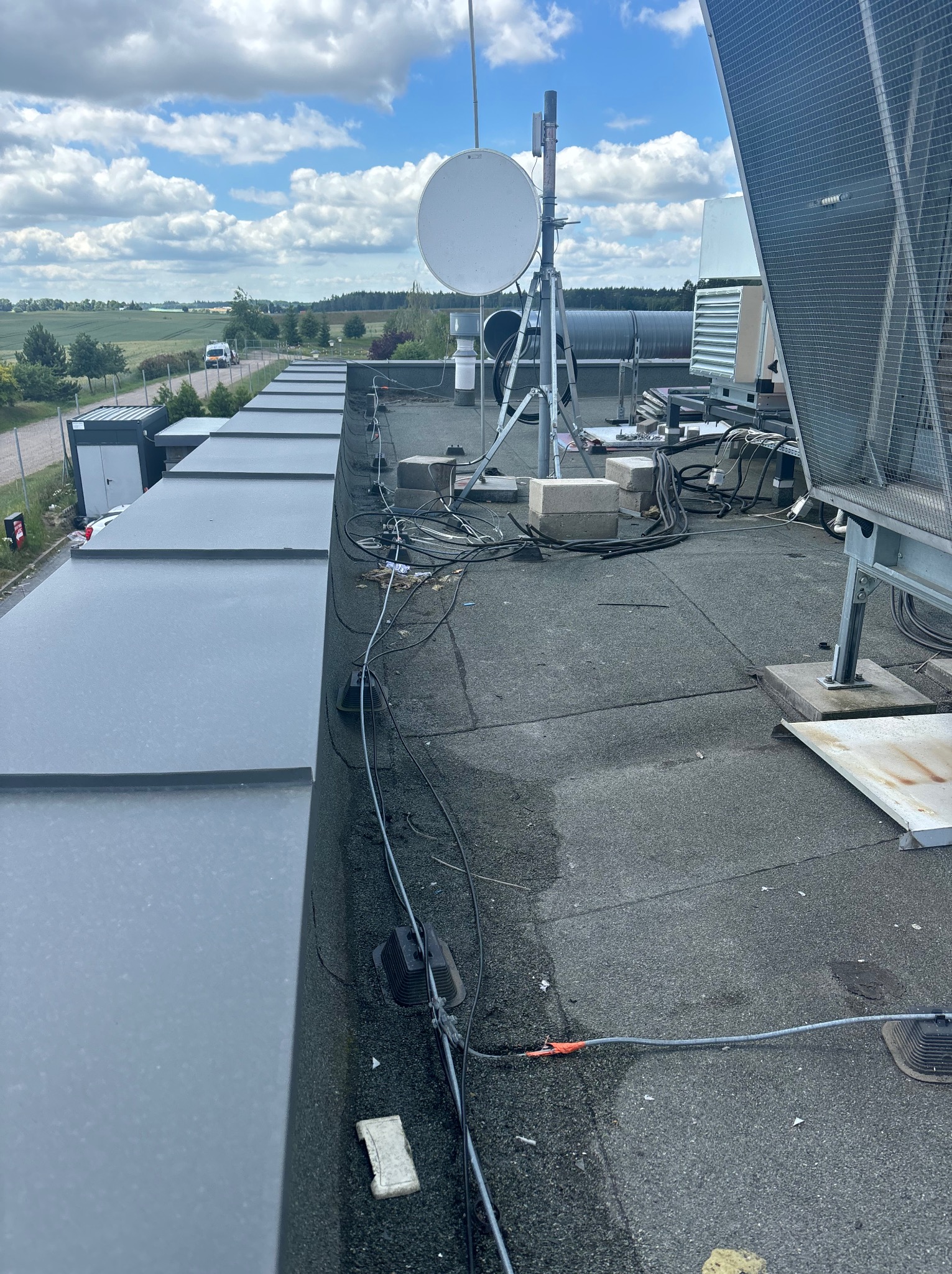 Płaski dach pokryty papą z widocznymi instalacjami telekomunikacyjnymi: anteny satelitarne, kable, oraz elementy mocujące. W tle widoczne budynki kontenerowe i krajobraz z polami i lasami...