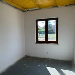 Wnętrze pokoju po pracach przygotowawczych w Lubięcinie: gładkie, białe ściany, okno z widokiem na ogród i surowy beton na podłodze. Sufit z żółtą izolacją.