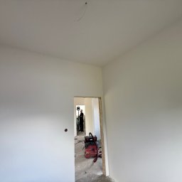 FAST-BUD Ireneusz Habura - Wnętrze po malowaniu na biało, widoczny pracownik z lampą i odkurzacz. Świeżo pomalowane ściany i sufit. Gładkie powierzchnie.