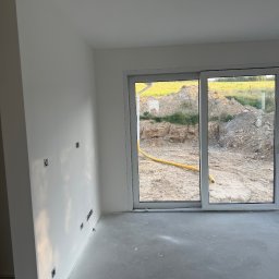 FAST-BUD Ireneusz Habura - Jasne wnętrze z gładkimi, białymi ścianami i szarą podłogą. Widoczne gniazdka elektryczne i okno z widokiem na plac budowy z żółtym wężem.