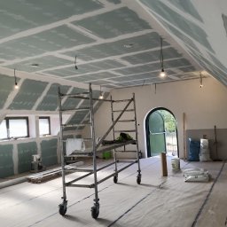 FAST-BUD Ireneusz Habura - Poddasze z oknami, częściowo pokryte płytami kartonowo-gipsowymi, z rozstawionym rusztowaniem i materiałami budowlanymi na podłodze, w trakcie prac wykończeniowych.