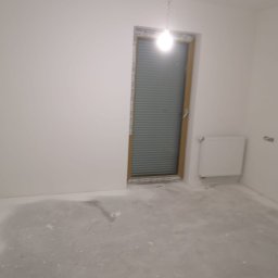 FAST-BUD Ireneusz Habura - Wnętrze pokoju po szpachlowaniu ścian i przygotowaniu podłoża. Widoczne gładkie, białe ściany, okno z żaluzjami i grzejnik. Podłoga w trakcie prac wykończeniowych.