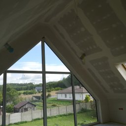 FAST-BUD Ireneusz Habura - Poddasze z oknem o nietypowym kształcie, ściany i sufit przygotowane pod malowanie, widoczne łączenia płyt gipsowych. W tle zieleń i zabudowa.