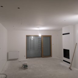 FAST-BUD Ireneusz Habura - Wnętrze po szpachlowaniu: białe ściany, okno z żaluzjami, grzejnik, obudowany kominek z czarnymi elementami. Na podłodze widoczne worki z zaprawą.