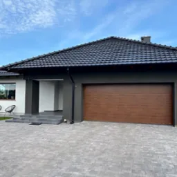 Nowoczesny dom jednorodzinny z grafitową elewacją, brązową bramą garażową i czarnym dachem z dachówki ceramicznej. Uwagę zwraca precyzyjnie ułożona kostka brukowa.