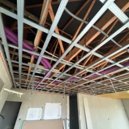 Montaż sufitu podwieszanego z widoczną konstrukcją metalową, rurami wentylacyjnymi i elementami drewnianymi w surowym wnętrzu z planami na ścianie.