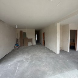 FAST-BUD Ireneusz Habura - Surowe wnętrze w trakcie prac: szare ściany, betonowa podłoga, widoczne kable i materiały budowlane. Przestrzeń przygotowana do dalszych prac wykończeniowych.