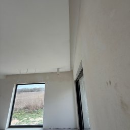 FAST-BUD Ireneusz Habura - Wnętrze w trakcie prac wykończeniowych: ściany przygotowane pod malowanie, widoczne przewody elektryczne, okno z widokiem na pole. Minimalistyczny styl.