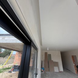 FAST-BUD Ireneusz Habura - Wnętrze w trakcie prac wykończeniowych: gładkie ściany i sufit, widoczne płyty karton-gips, okno z czarną ramą. Na zewnątrz widoczny teren budowy.