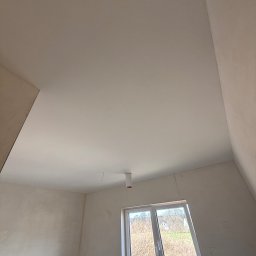 FAST-BUD Ireneusz Habura - Wnętrze poddasza po szpachlowaniu, widoczne gładkie ściany i sufit, okno z widokiem na zewnątrz. Świeżo wykończone powierzchnie gotowe do dalszych prac.