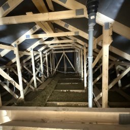 FAST-BUD Ireneusz Habura - Szkielet dachu z izolacją z wełny mineralnej, widok z dołu. Elementy drewniane łączone metalowymi płytkami. Rura wentylacyjna pionowo w kadrze.