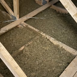 FAST-BUD Ireneusz Habura - Ocieplenie poddasza wełną mineralną, widoczne drewniane elementy konstrukcyjne i biały przewód elektryczny. Ujęcie z góry, wypełnienie przestrzeni izolacją.