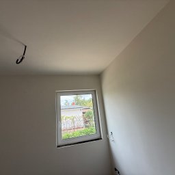 FAST-BUD Ireneusz Habura - Jasne wnętrze po remoncie: świeżo pomalowane ściany w neutralnym odcieniu, okno z widokiem na zieleń, wystające kable elektryczne. Minimalizm i czystość.