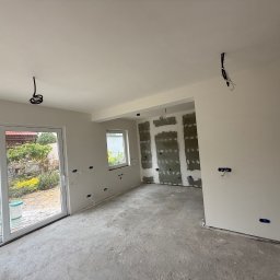 FAST-BUD Ireneusz Habura - Wnętrze w trakcie prac: ściany z instalacją elektryczną, częściowo obłożone płytami, surowa podłoga, widok na ogród przez okno. Etap przygotowania do dalszych prac.