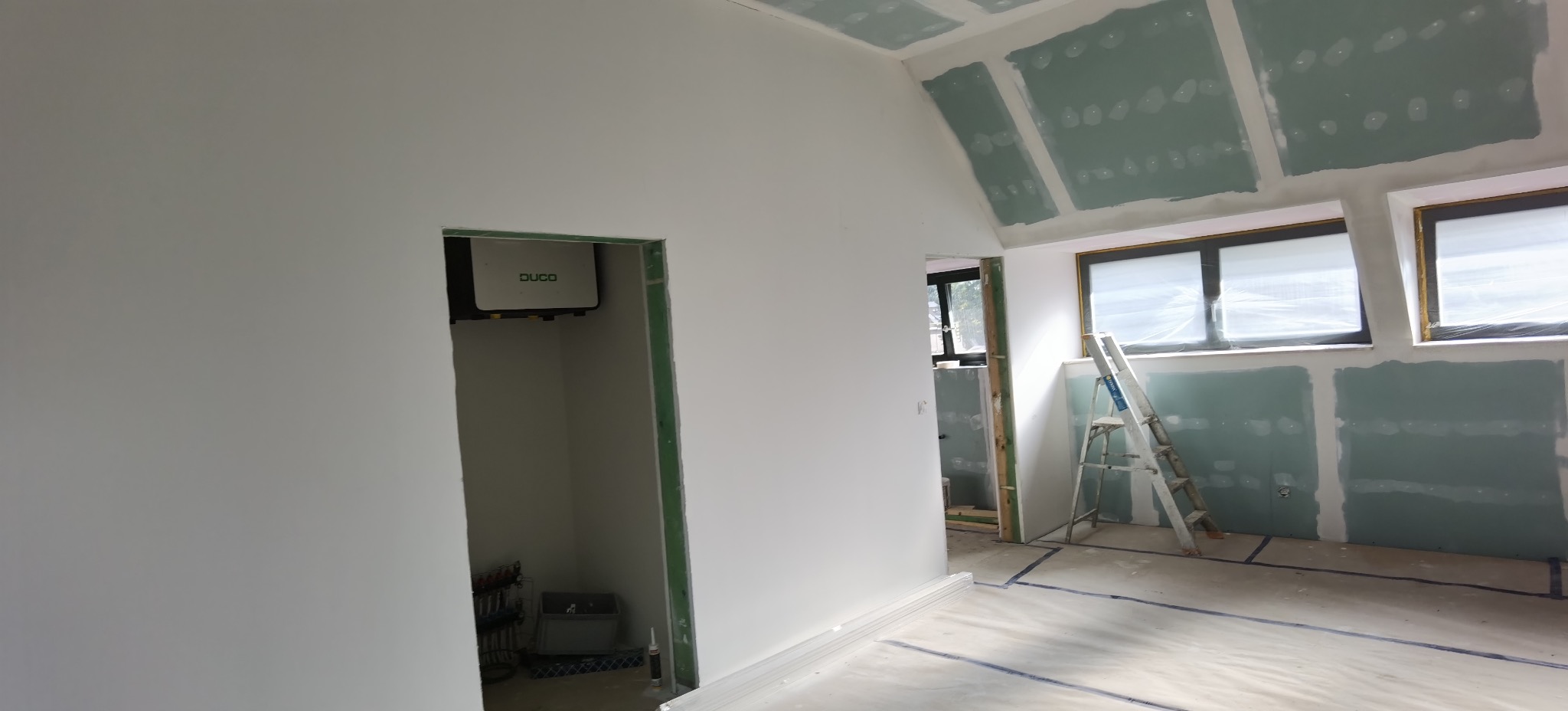 Wnętrze w trakcie prac wykończeniowych: ściany pokryte płytami gipsowo-kartonowymi, widoczna drabina, okna zabezpieczone folią, przygotowane do dalszych prac.