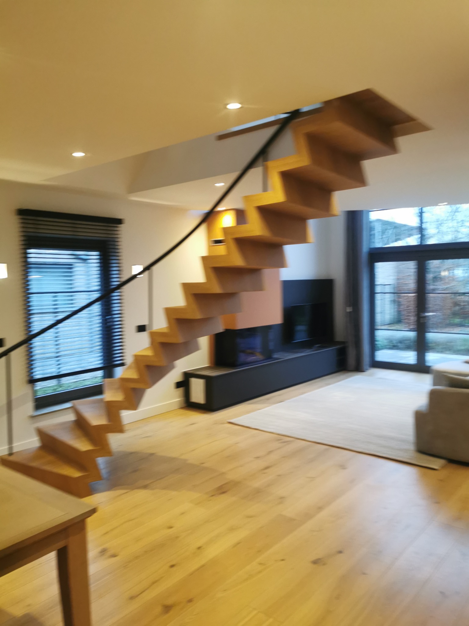 Nowoczesne, drewniane schody wiszące z czarną poręczą w jasnym wnętrzu z parkietem, oknem z żaluzjami i dużym oknem balkonowym.