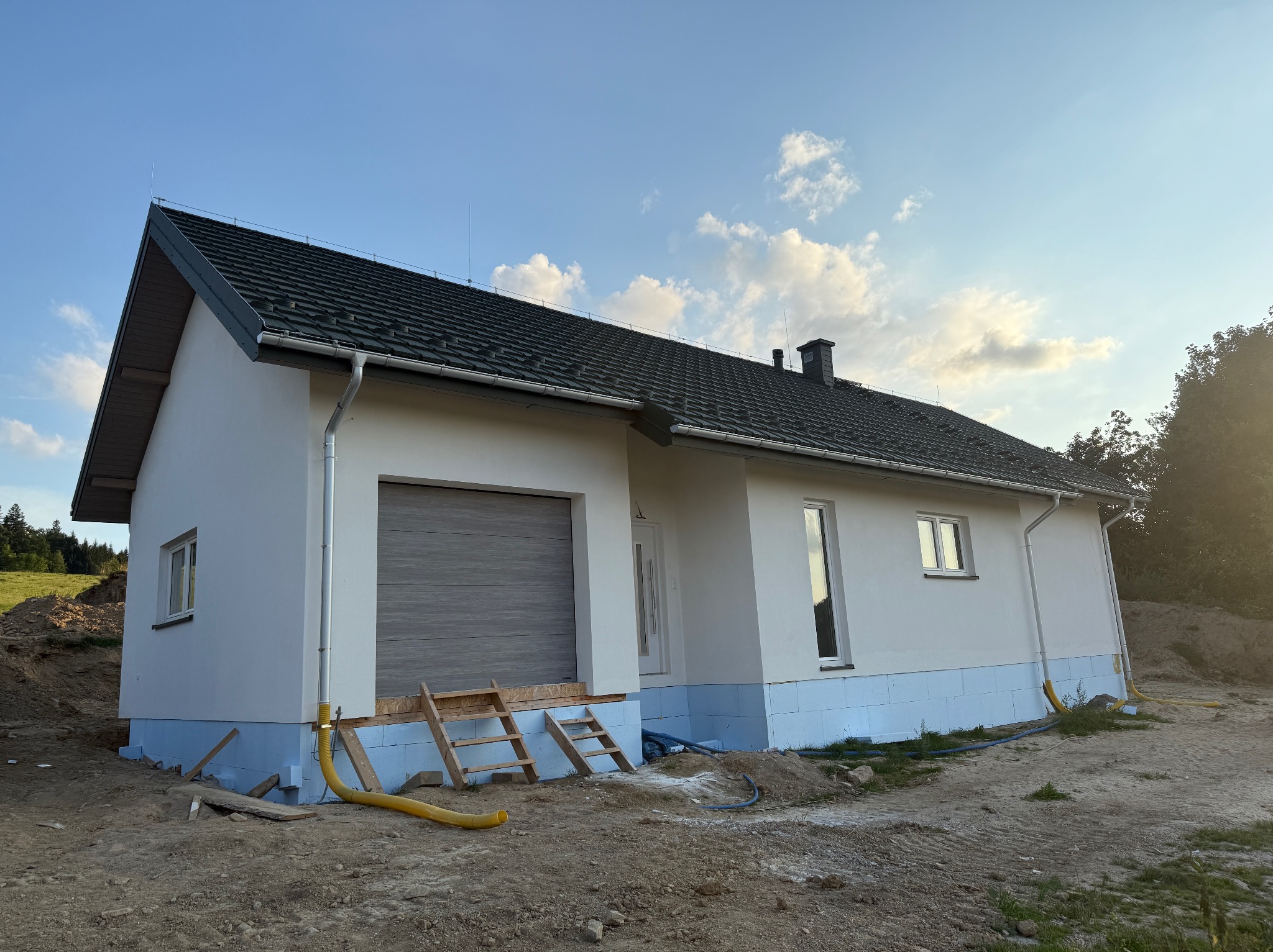 Biały dom jednorodzinny z szarą dachówką i garażem, ocieplony przy gruncie niebieskim styropianem, widoczny na tle błękitnego nieba z chmurami i zieleni.