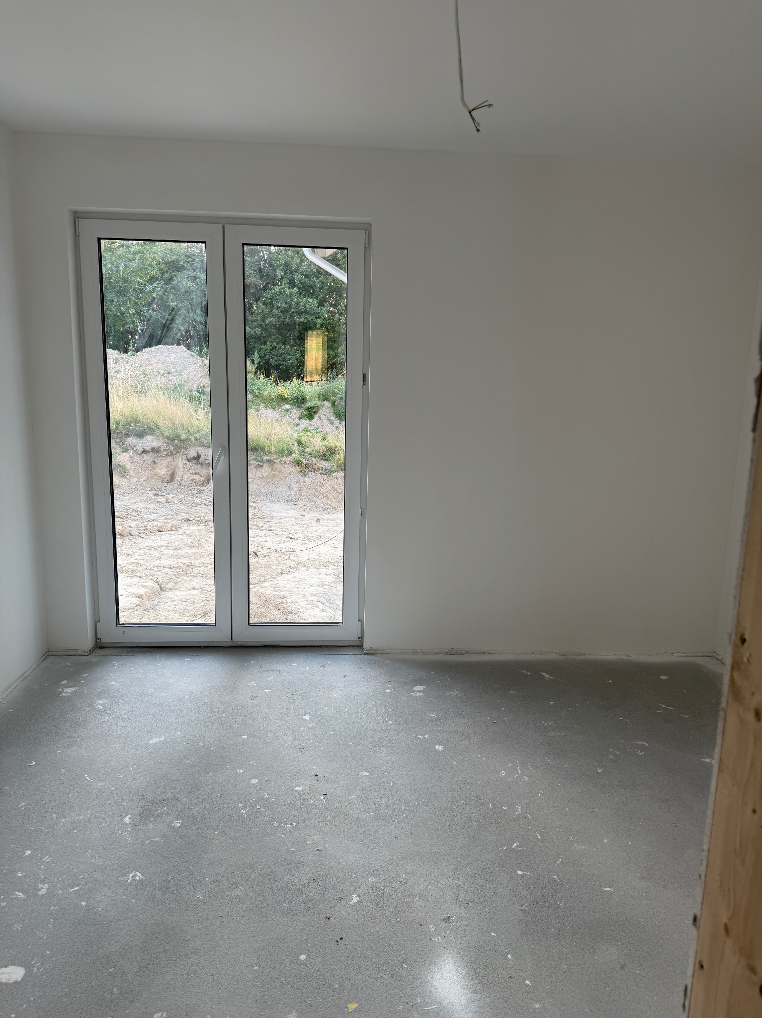 Surowe wnętrze pokoju z białymi ścianami, betonową podłogą i widokiem na teren zewnętrzny przez dwuskrzydłowe drzwi balkonowe. Widoczne niedociągnięcia na podłodze.
