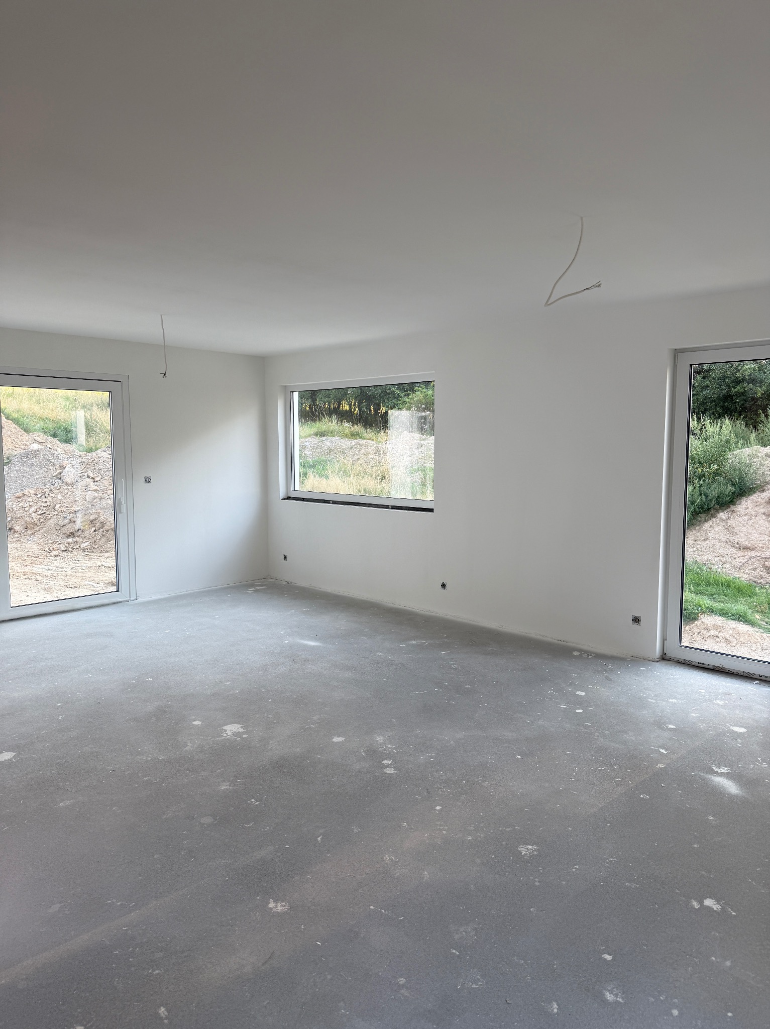 Surowe wnętrze z oknami w Lubięcinie, ściany świeżo pomalowane na biało, podłoga zacierana, przygotowane do dalszych prac wykończeniowych. Widok na zewnątrz.