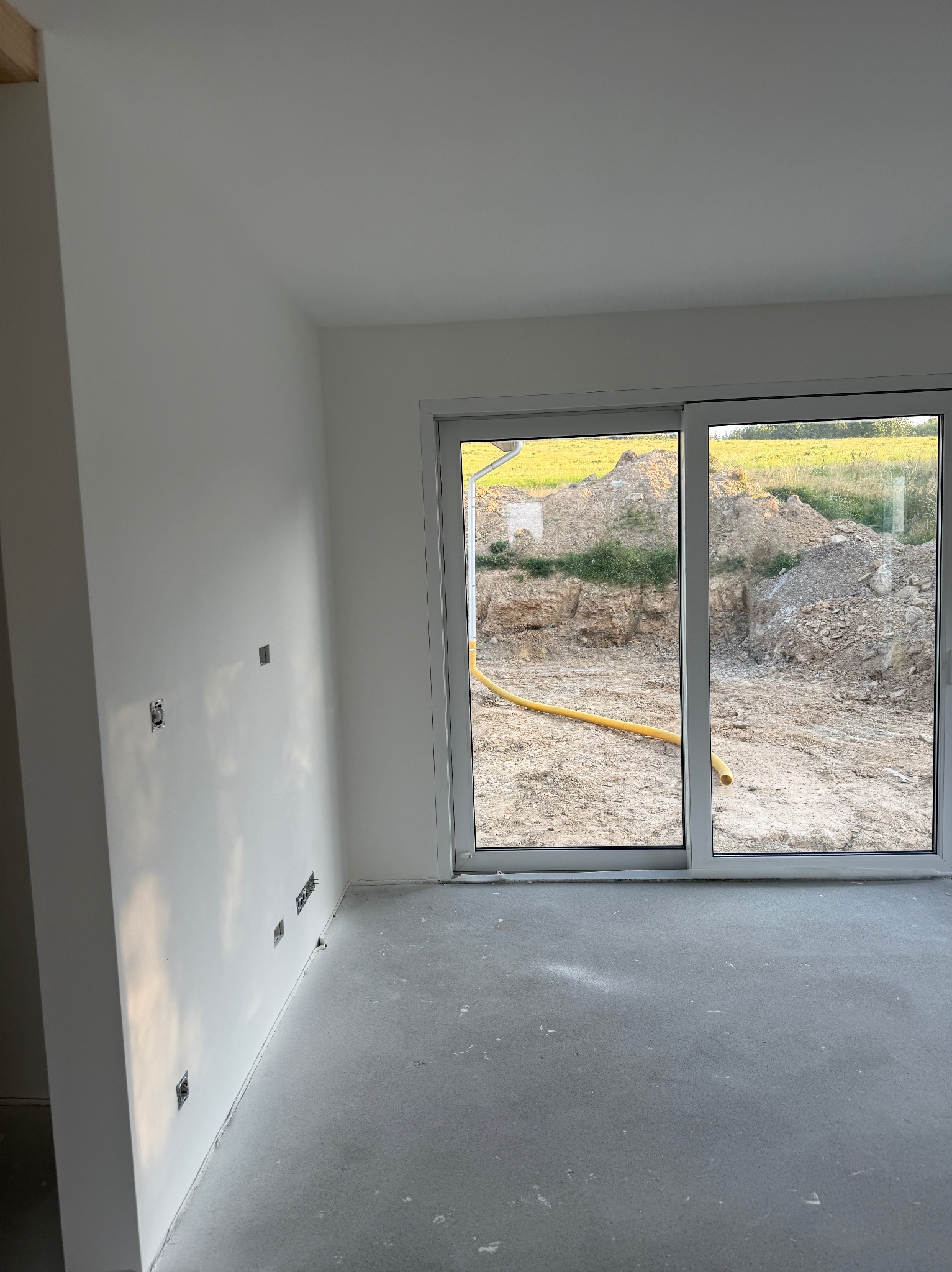 Jasne wnętrze z gładkimi, białymi ścianami i szarą podłogą. Widoczne gniazdka elektryczne i okno z widokiem na plac budowy z żółtym wężem.