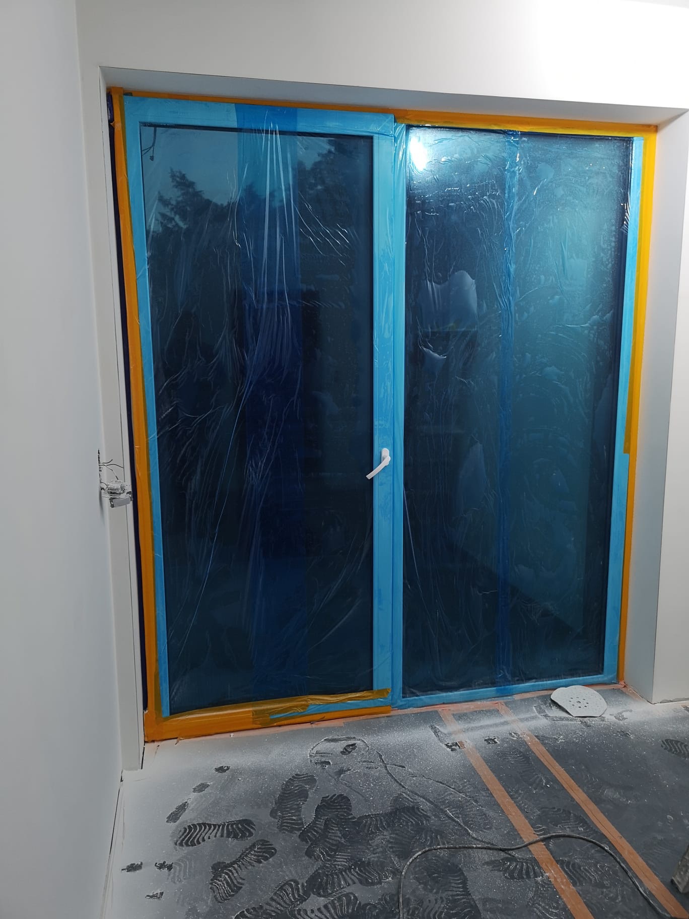 Okna zabezpieczone folią i taśmą malarską przed pracami wykończeniowymi. Widoczne ślady pyłu na podłodze. Przygotowanie do prac remontowych.