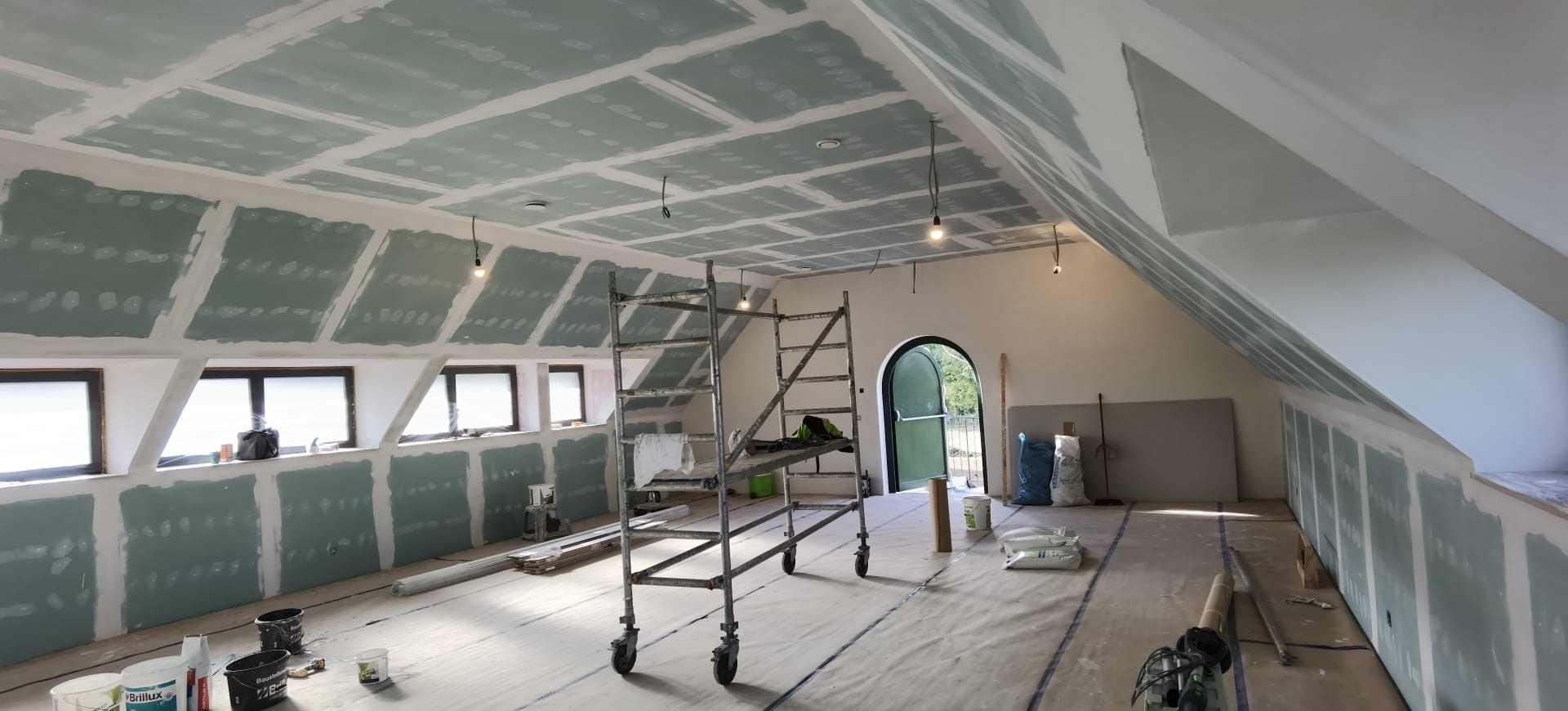 Poddasze z oknami, częściowo pokryte płytami kartonowo-gipsowymi, z rozstawionym rusztowaniem i materiałami budowlanymi na podłodze, w trakcie prac wykończeniowych.