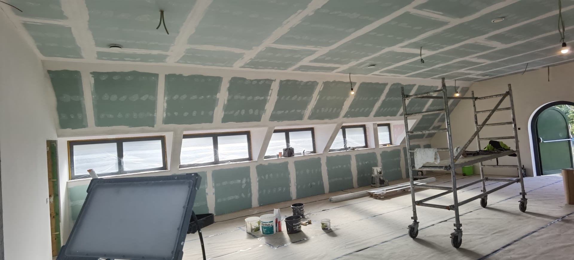 Poddasze z oknami dachowymi w trakcie montażu płyt kartonowo-gipsowych, widoczne łączenia płyt, rusztowanie i narzędzia. Prace wykończeniowe w jasnym pomieszczeniu.