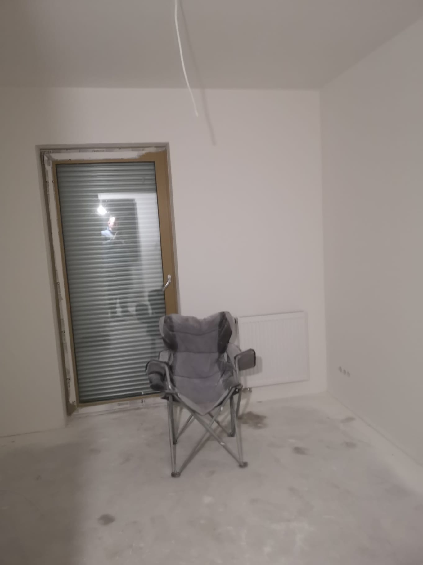 Wnętrze pokoju w trakcie prac wykończeniowych: białe ściany, okno z żaluzjami, grzejnik i szare krzesło turystyczne na środku. Widoczne ślady po pracach na podłodze.