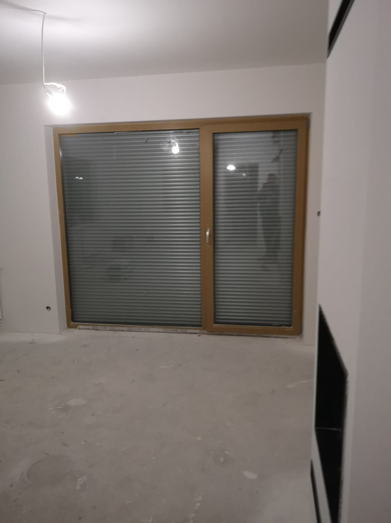 Wnętrze pokoju przed wykończeniem: surowe ściany, betonowa podłoga, okno z żaluzjami i wisząca żarówka. Widoczny fragment zabudowy meblowej po prawej.