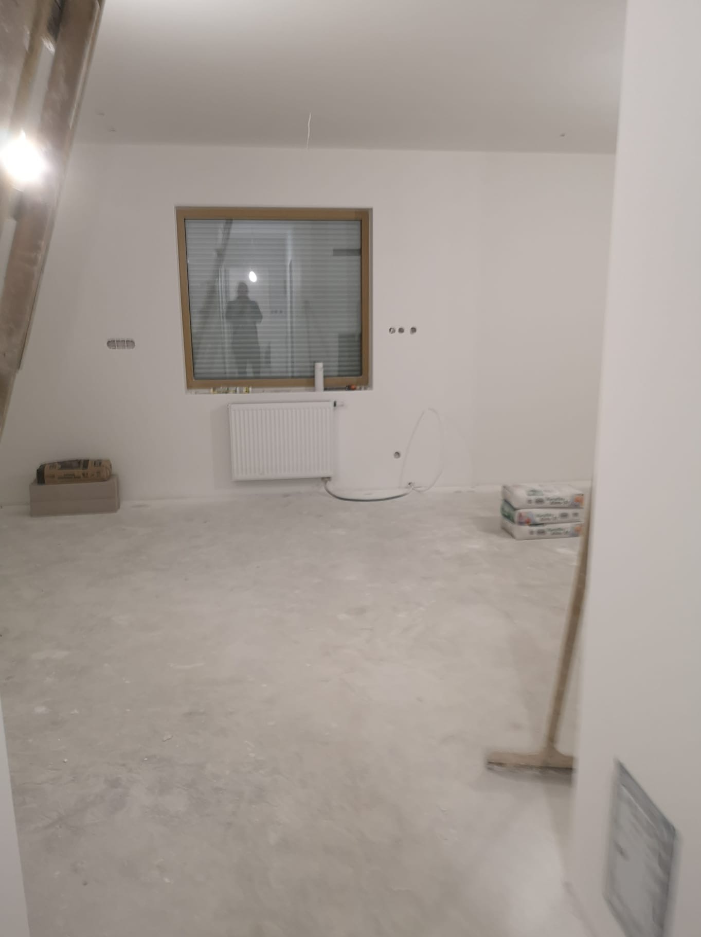 Wnętrze pokoju po wylewce, przygotowane do dalszych prac wykończeniowych. Widoczne worki z materiałami, okno, grzejnik i ślady po szpachlowaniu na ścianach.