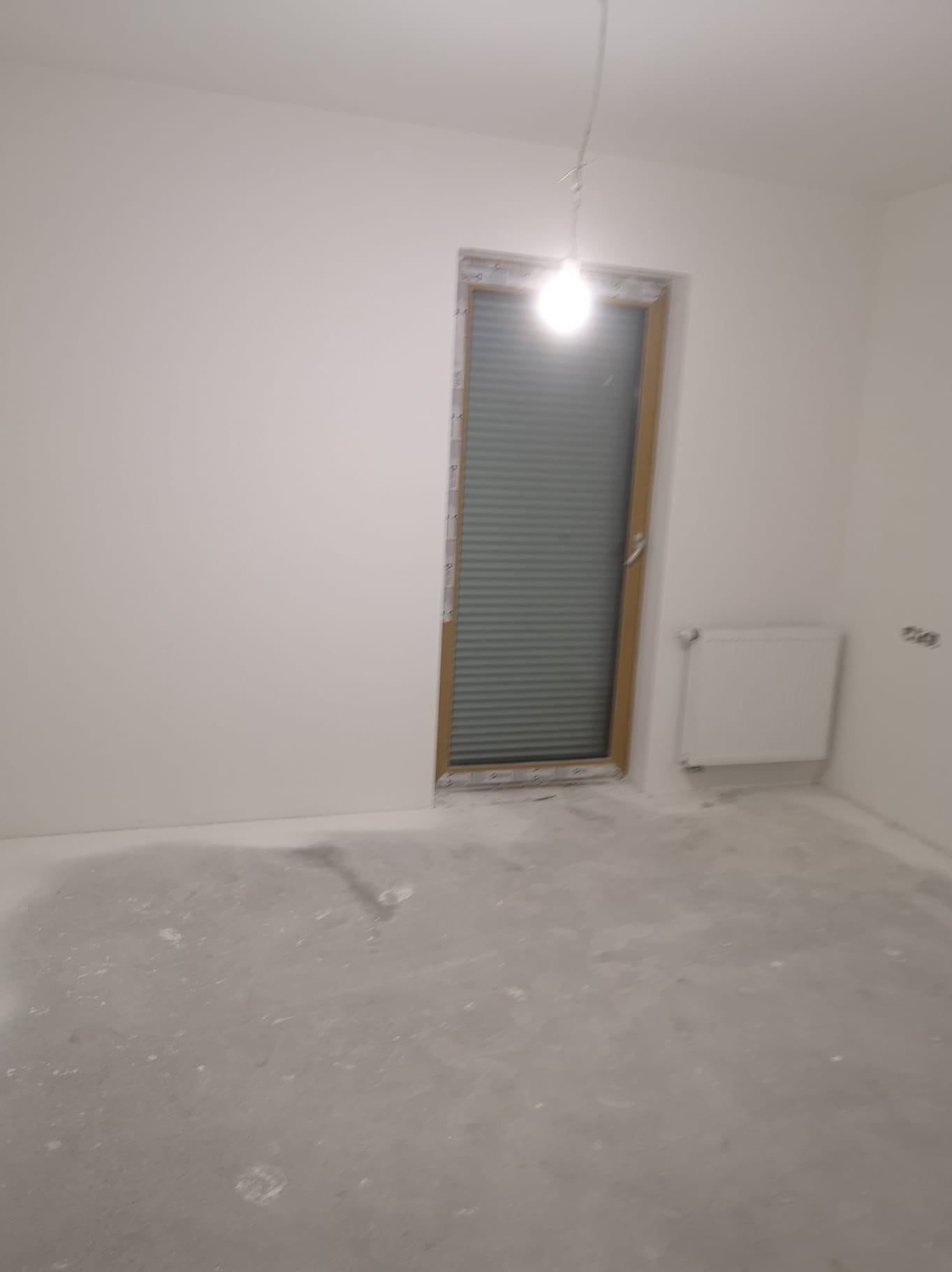 Wnętrze pokoju po szpachlowaniu ścian i przygotowaniu podłoża. Widoczne gładkie, białe ściany, okno z żaluzjami i grzejnik. Podłoga w trakcie prac wykończeniowych.