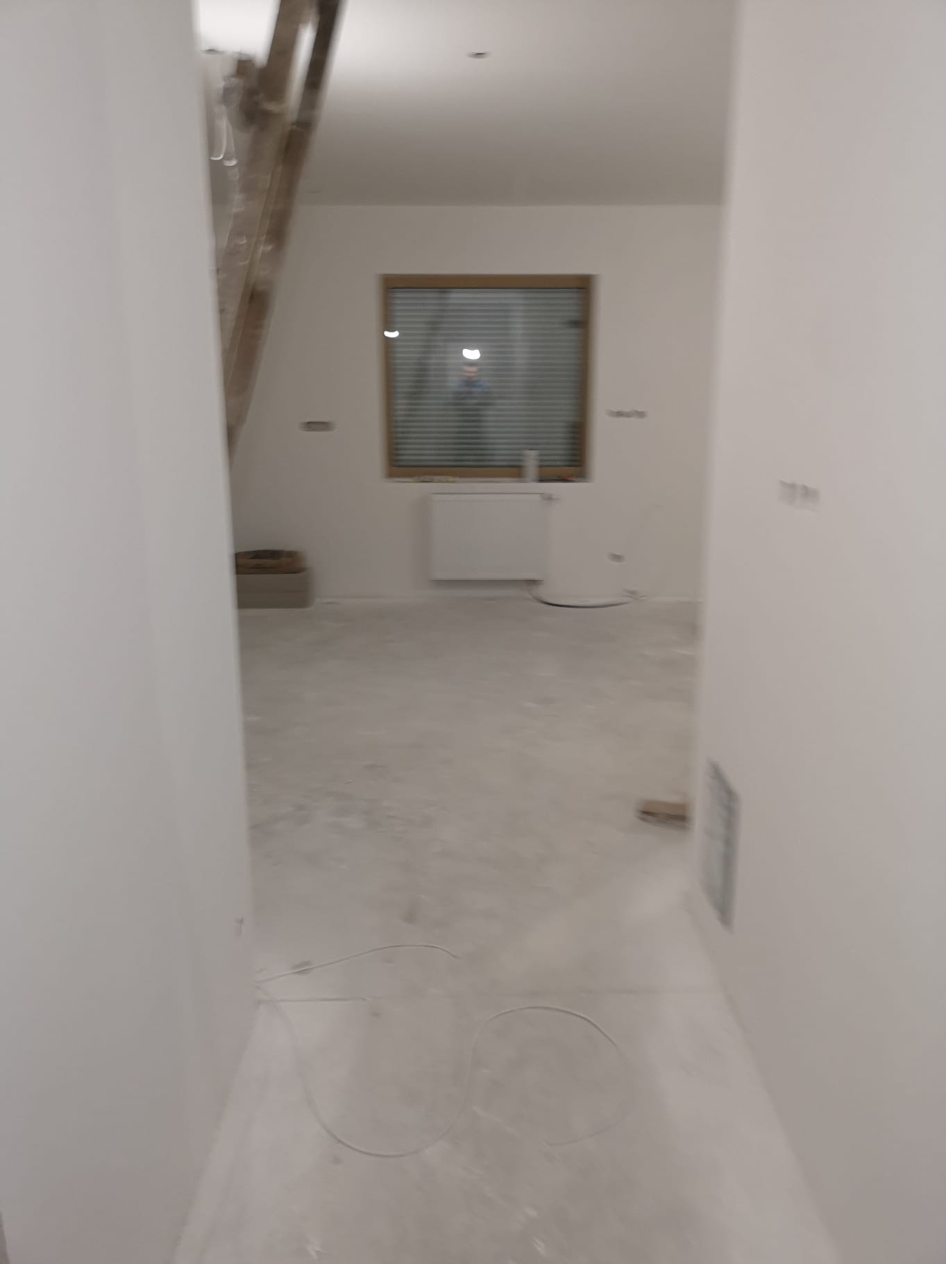 Wnętrze w trakcie prac wykończeniowych: białe ściany, surowa podłoga, okno z żaluzjami. Widoczne przewody elektryczne na podłodze, schody w tle.