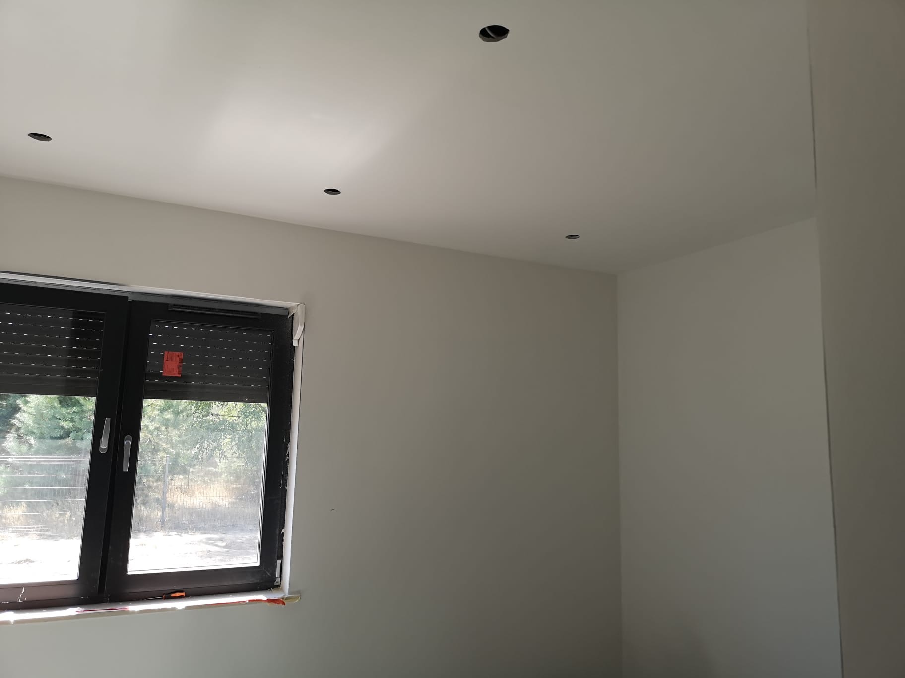 Wnętrze pokoju po remoncie z gładkimi, pomalowanymi na szaro ścianami i sufitem. Widoczne otwory na oświetlenie sufitowe i okno z roletami.