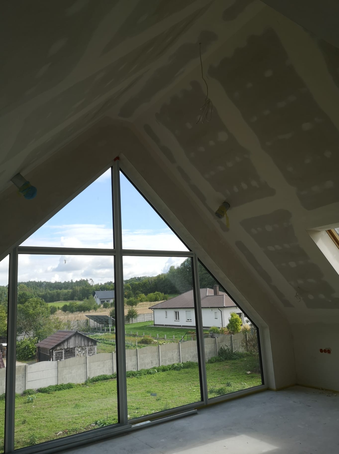 Poddasze z oknem o nietypowym kształcie, ściany i sufit przygotowane pod malowanie, widoczne łączenia płyt gipsowych. W tle zieleń i zabudowa.