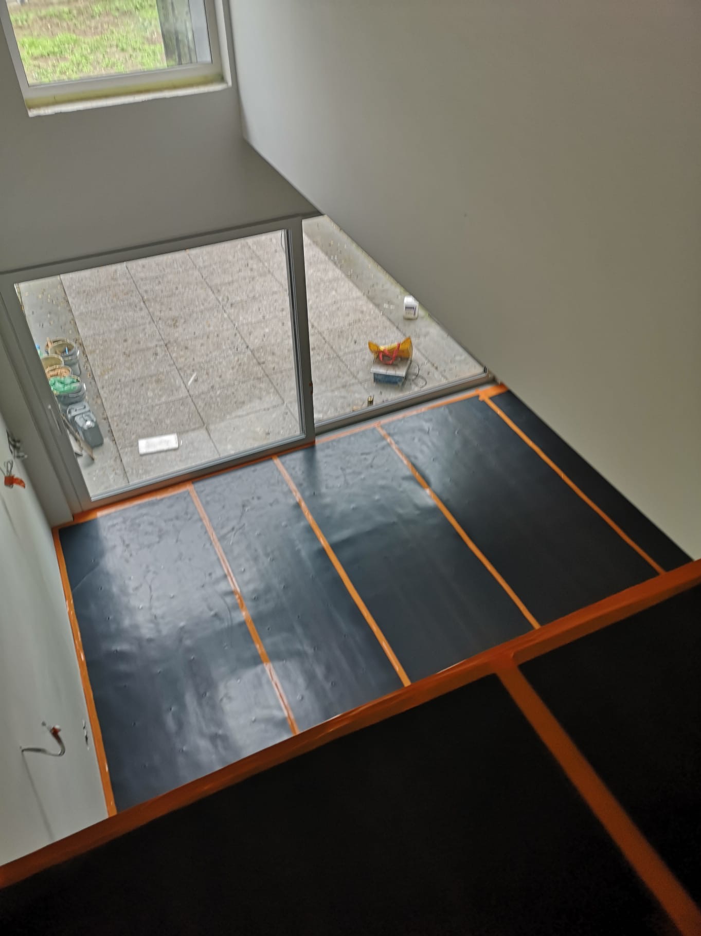 Czarna folia grzewcza z pomarańczowymi paskami, rozłożona na podłodze w pomieszczeniu z dużym oknem, widok z góry. Etap prac instalacyjnych.