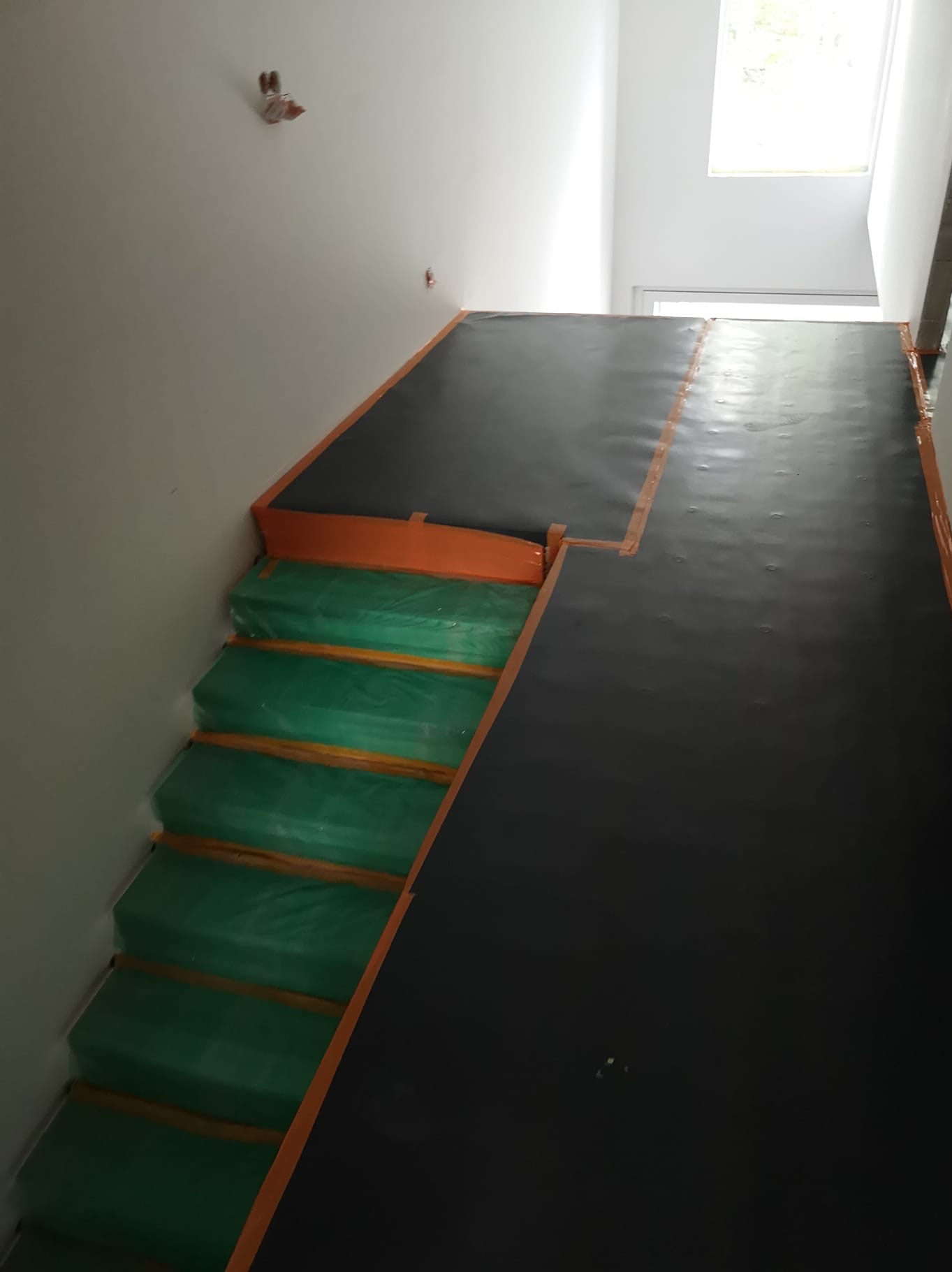 Schody i podłoga zabezpieczone folią i taśmą malarską przed pracami wykończeniowymi. Widoczne przygotowania do malowania lub innego rodzaju prac.