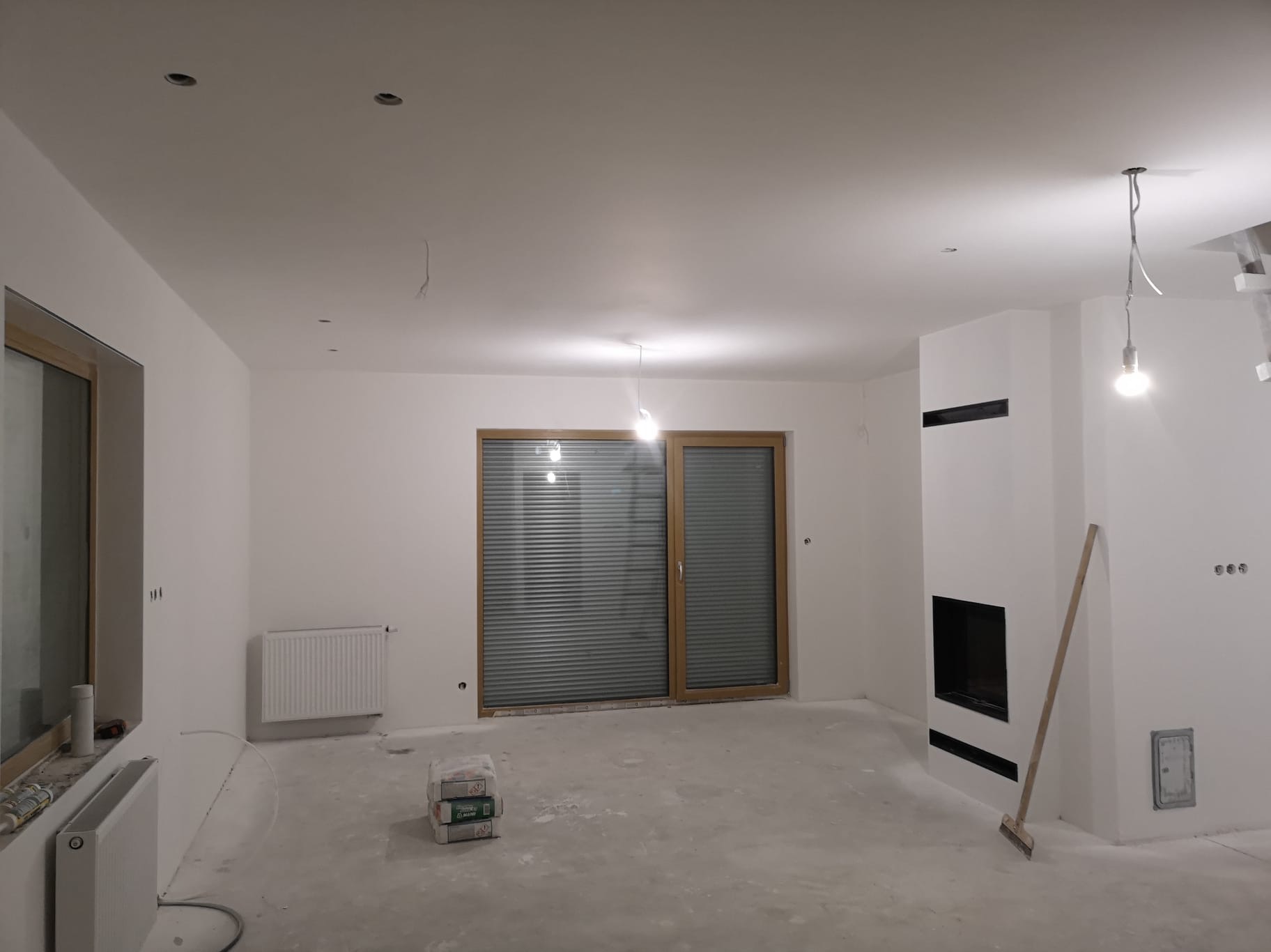 Wnętrze po szpachlowaniu: białe ściany, okno z żaluzjami, grzejnik, obudowany kominek z czarnymi elementami. Na podłodze widoczne worki z zaprawą.