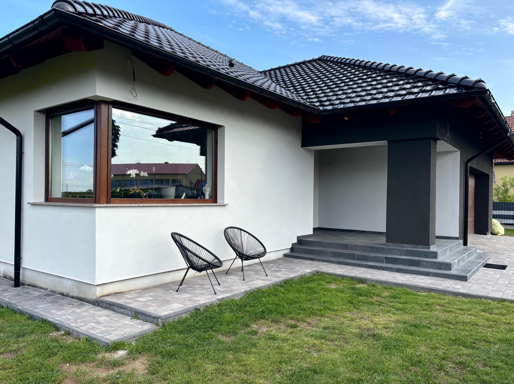 Biały dom jednorodzinny z czarnym dachem, widok z zewnątrz. Elewacja wykończona, z widocznymi dwoma krzesłami na trawniku. Nowoczesny styl.