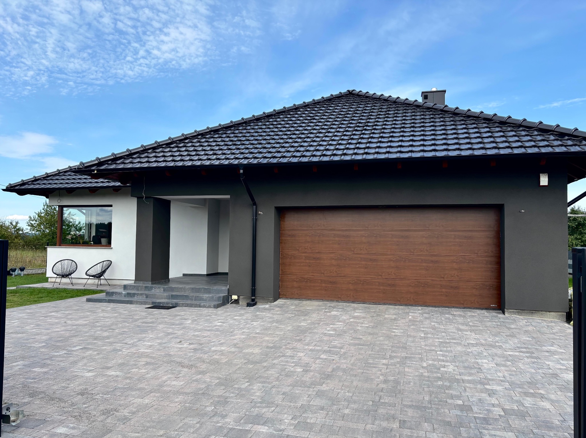 Nowoczesny dom jednorodzinny z grafitową elewacją, brązową bramą garażową i czarnym dachem z dachówki ceramicznej. Uwagę zwraca precyzyjnie ułożona kostka brukowa.