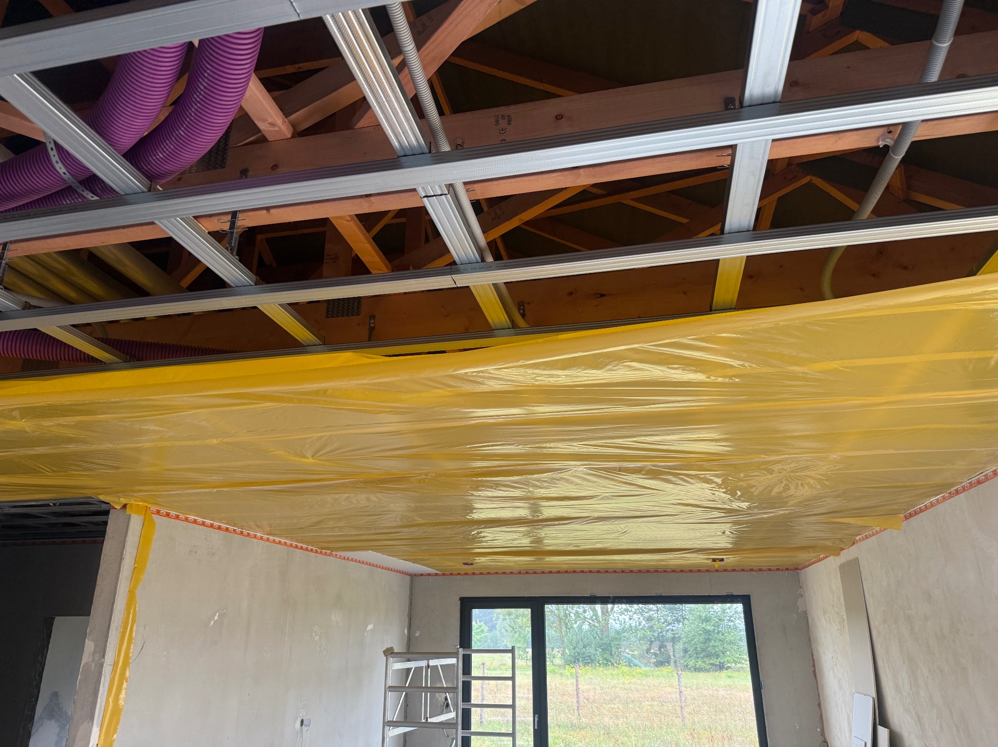 Montaż sufitu podwieszanego z widoczną konstrukcją metalową, izolacją w kolorze żółtym i elementami instalacji wentylacyjnej. Ściany w stanie surowym, okno z widokiem na zieleń.