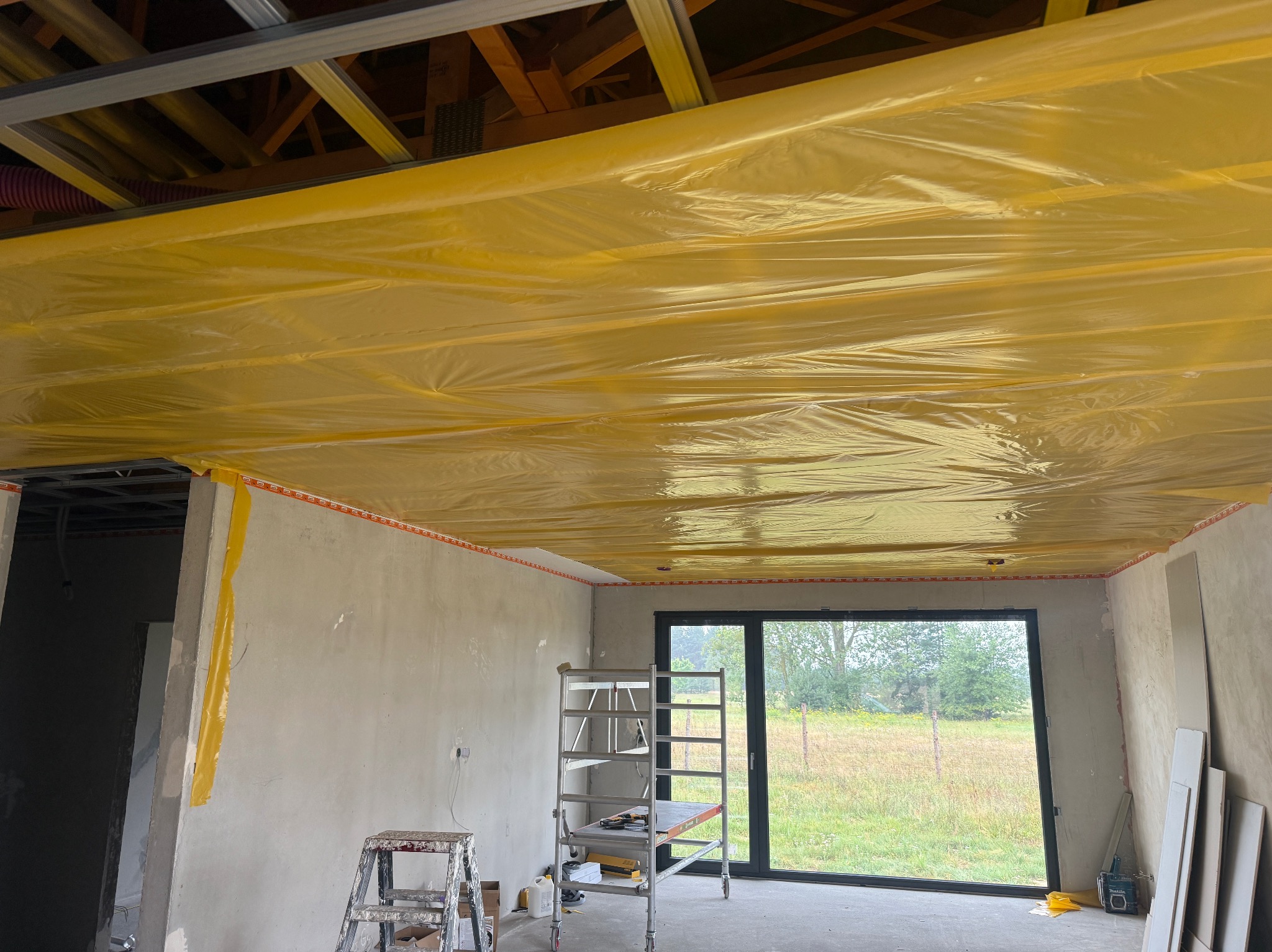 Żółta folia paroizolacyjna na suficie podwieszanym, widoczne profile i drabina. Ściany surowe, przygotowane do dalszych prac. Duże okno z widokiem na zieleń.